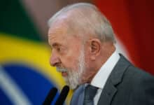Um homem de cabelo grisalho e barba branca está falando em um microfone. Ele usa um terno cinza e uma gravata azul. Ao fundo, há uma bandeira do Brasil e outras bandeiras desfocadas. O homem parece estar em um evento oficial, possivelmente dando um discurso.