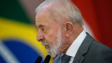 Um homem de cabelo grisalho e barba branca está falando em um microfone. Ele usa um terno cinza e uma gravata azul. Ao fundo, há uma bandeira do Brasil e outras bandeiras desfocadas. O homem parece estar em um evento oficial, possivelmente dando um discurso.