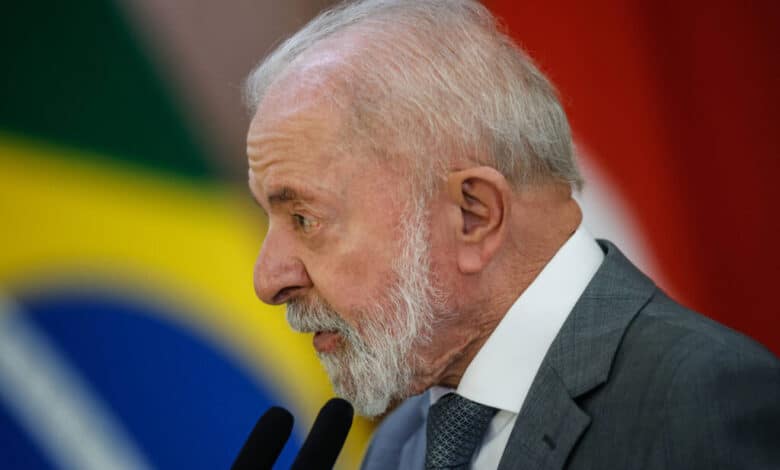 Um homem de cabelo grisalho e barba branca está falando em um microfone. Ele usa um terno cinza e uma gravata azul. Ao fundo, há uma bandeira do Brasil e outras bandeiras desfocadas. O homem parece estar em um evento oficial, possivelmente dando um discurso.