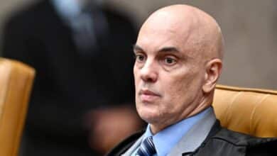 Homem de meia idade, calvo, vestindo terno escuro, camisa azul clara e gravata listrada, aparece sentado em uma poltrona, com fundo desfocado