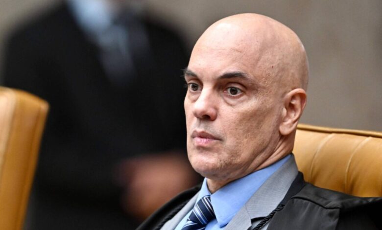 Homem de meia idade, calvo, vestindo terno escuro, camisa azul clara e gravata listrada, aparece sentado em uma poltrona, com fundo desfocado