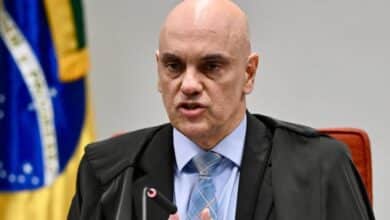 Um homem careca, vestido com uma toga preta e uma camisa azul, está falando em uma audiência. Ele gesticula com a mão direita enquanto segura um microfone com a mão esquerda. Ao fundo, há uma bandeira do Brasil visível, com as cores verde, amarelo e azul.