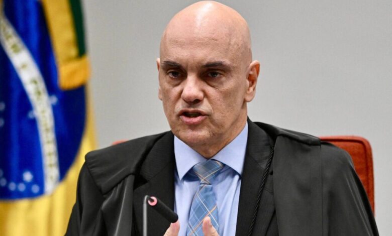 Um homem careca, vestido com uma toga preta e uma camisa azul, está falando em uma audiência. Ele gesticula com a mão direita enquanto segura um microfone com a mão esquerda. Ao fundo, há uma bandeira do Brasil visível, com as cores verde, amarelo e azul.