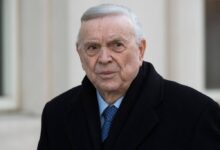 Um homem idoso com cabelo grisalho e pele clara, usando um casaco preto e uma gravata azul. Ele está olhando para a câmera com uma expressão séria, em um ambiente externo com paredes claras ao fundo.
