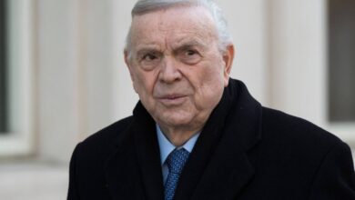 Um homem idoso com cabelo grisalho e pele clara, usando um casaco preto e uma gravata azul. Ele está olhando para a câmera com uma expressão séria, em um ambiente externo com paredes claras ao fundo.