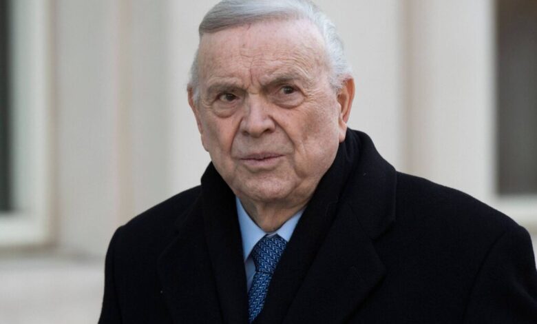 Um homem idoso com cabelo grisalho e pele clara, usando um casaco preto e uma gravata azul. Ele está olhando para a câmera com uma expressão séria, em um ambiente externo com paredes claras ao fundo.