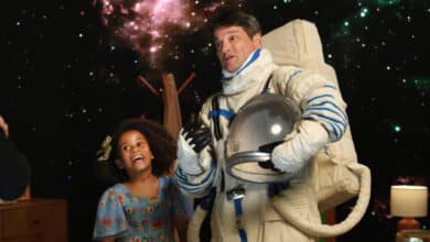A imagem mostra um homem vestido de astronauta, usando um traje espacial completo e um capacete, ao lado de uma menina sorridente. A menina está vestindo um vestido colorido e parece estar se divertindo. O fundo da imagem é uma representação do espaço, com estrelas e nebulosas visíveis. No ambiente, há móveis como uma cômoda e uma lâmpada, sugerindo que a cena pode ser em um estúdio ou quarto.