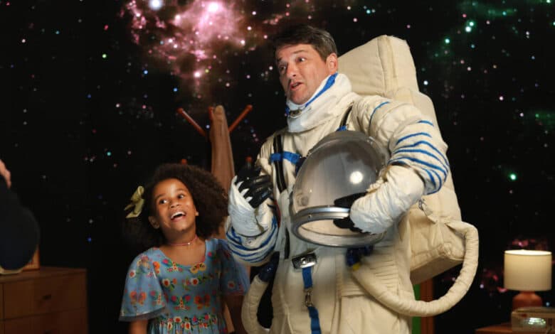 A imagem mostra um homem vestido de astronauta, usando um traje espacial completo e um capacete, ao lado de uma menina sorridente. A menina está vestindo um vestido colorido e parece estar se divertindo. O fundo da imagem é uma representação do espaço, com estrelas e nebulosas visíveis. No ambiente, há móveis como uma cômoda e uma lâmpada, sugerindo que a cena pode ser em um estúdio ou quarto.