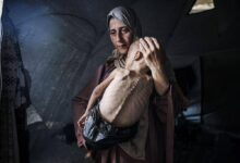 A imagem mostra uma mulher com um lenço na cabeça segurando uma criança magra em seus braços. A criança parece estar em estado de desnutrição, com o corpo visivelmente esquelético. O ambiente ao redor é de uma tenda, com luz suave entrando por uma abertura. A expressão da mulher é de preocupação e tristeza.