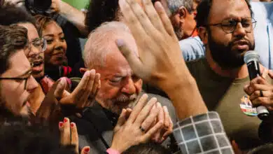 A imagem mostra um grupo de pessoas em um evento político, com destaque para um homem de cabelos grisalhos e barba, que parece emocionado, com os olhos fechados e as mãos unidas em um gesto de oração ou agradecimento. Ao seu redor, várias mãos estão levantadas, algumas tocando-o, enquanto outras seguram microfones e câmeras. O ambiente é de intensa interação e apoio emocional.