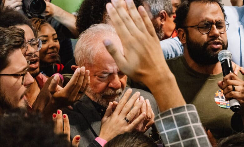 A imagem mostra um grupo de pessoas em um evento político, com destaque para um homem de cabelos grisalhos e barba, que parece emocionado, com os olhos fechados e as mãos unidas em um gesto de oração ou agradecimento. Ao seu redor, várias mãos estão levantadas, algumas tocando-o, enquanto outras seguram microfones e câmeras. O ambiente é de intensa interação e apoio emocional.