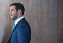 Um homem está posicionado de perfil, olhando para a direita. Ele tem cabelo curto e barba bem aparada. Está vestindo um terno azul escuro e uma camisa branca com uma gravata azul clara. O fundo é uma parede de madeira clara, criando um contraste com sua roupa.