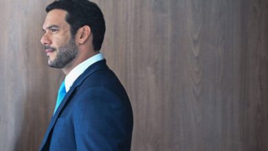 Um homem está posicionado de perfil, olhando para a direita. Ele tem cabelo curto e barba bem aparada. Está vestindo um terno azul escuro e uma camisa branca com uma gravata azul clara. O fundo é uma parede de madeira clara, criando um contraste com sua roupa.