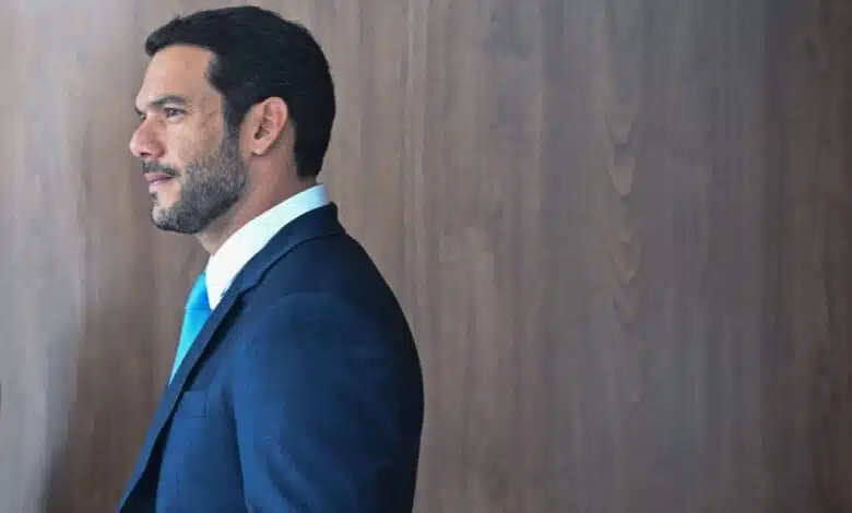 Um homem está posicionado de perfil, olhando para a direita. Ele tem cabelo curto e barba bem aparada. Está vestindo um terno azul escuro e uma camisa branca com uma gravata azul clara. O fundo é uma parede de madeira clara, criando um contraste com sua roupa.