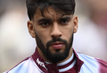 Um jovem jogador de futebol com cabelo escuro e barba, vestindo uma camisa de treino com as cores marrom e azul. Ele está olhando diretamente para a câmera com uma expressão séria. O fundo é desfocado, sugerindo um ambiente de estádio.