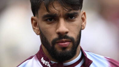 Um jovem jogador de futebol com cabelo escuro e barba, vestindo uma camisa de treino com as cores marrom e azul. Ele está olhando diretamente para a câmera com uma expressão séria. O fundo é desfocado, sugerindo um ambiente de estádio.