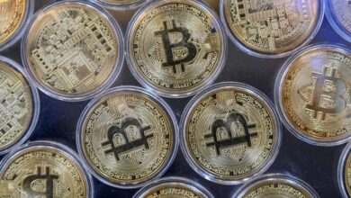 A imagem mostra várias moedas de Bitcoin dispostas em uma superfície. As moedas são de cor dourada e apresentam o símbolo do Bitcoin em destaque, com detalhes de circuitos eletrônicos ao fundo. Elas estão organizadas em um padrão circular, com algumas moedas parcialmente sobrepostas.