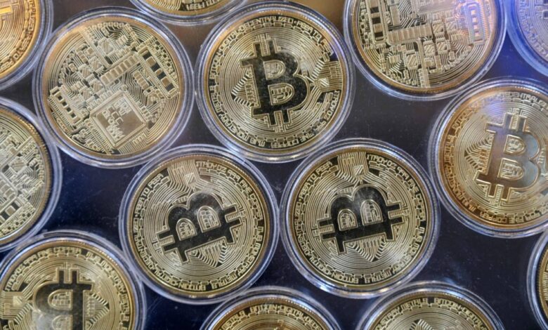 A imagem mostra várias moedas de Bitcoin dispostas em uma superfície. As moedas são de cor dourada e apresentam o símbolo do Bitcoin em destaque, com detalhes de circuitos eletrônicos ao fundo. Elas estão organizadas em um padrão circular, com algumas moedas parcialmente sobrepostas.