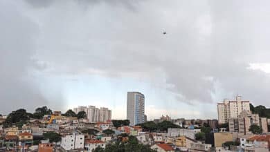 A imagem mostra uma vista panorâmica de uma área urbana, com prédios e casas. O céu está nublado, com nuvens escuras, sugerindo a possibilidade de chuva. Há um edifício alto em destaque no centro da imagem, cercado por construções menores. No fundo, é possível ver mais prédios e uma área verde.