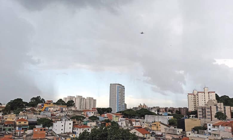 A imagem mostra uma vista panorâmica de uma área urbana, com prédios e casas. O céu está nublado, com nuvens escuras, sugerindo a possibilidade de chuva. Há um edifício alto em destaque no centro da imagem, cercado por construções menores. No fundo, é possível ver mais prédios e uma área verde.