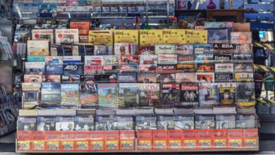 A imagem mostra uma banca de jornal com uma variedade de revistas e jornais expostos. A banca está organizada em prateleiras, com capas visíveis de diferentes publicações. À direita, uma mulher está de pé, usando uma máscara facial. O ambiente é iluminado e a banca é coberta por uma lona azul na parte superior.