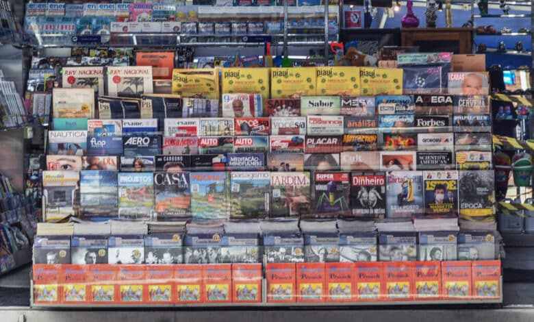 A imagem mostra uma banca de jornal com uma variedade de revistas e jornais expostos. A banca está organizada em prateleiras, com capas visíveis de diferentes publicações. À direita, uma mulher está de pé, usando uma máscara facial. O ambiente é iluminado e a banca é coberta por uma lona azul na parte superior.
