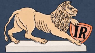 A imagem apresenta um leão em posição de alerta, com uma pata apoiada em um escudo. O leão é de cor clara, enquanto o escudo é laranja com as letras