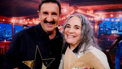 Na imagem, um homem e uma mulher estão sorrindo juntos em um ambiente iluminado. O homem, com bigode e cabelo curto, segura um troféu em forma de estrela. A mulher, com cabelo longo e grisalho, está segurando um buquê de flores coloridas. Ao fundo, há uma decoração com luzes e uma vista da cidade.