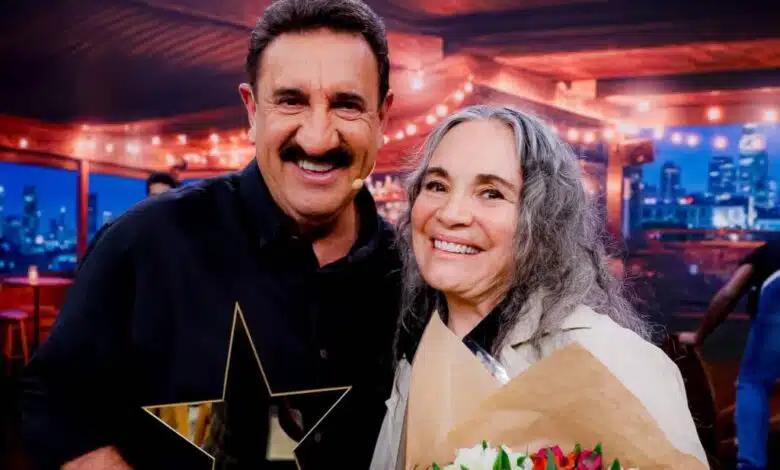 Na imagem, um homem e uma mulher estão sorrindo juntos em um ambiente iluminado. O homem, com bigode e cabelo curto, segura um troféu em forma de estrela. A mulher, com cabelo longo e grisalho, está segurando um buquê de flores coloridas. Ao fundo, há uma decoração com luzes e uma vista da cidade.