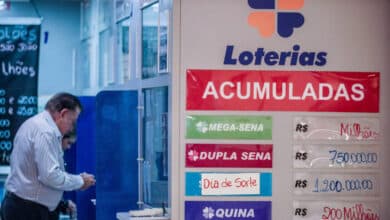 A imagem mostra um ambiente de loteria, com um homem em pé ao lado de um balcão. Na parede, há um cartaz grande com a palavra