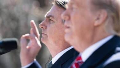 A imagem mostra dois homens em um evento ao ar livre. O homem à esquerda está de perfil, com a mão levantada, enquanto o homem à direita está em foco, mas parcialmente desfocado. Ambos parecem estar em um momento de discurso ou apresentação, com microfones à frente. O fundo é desfocado, sugerindo um ambiente de conferência ou coletiva de imprensa.