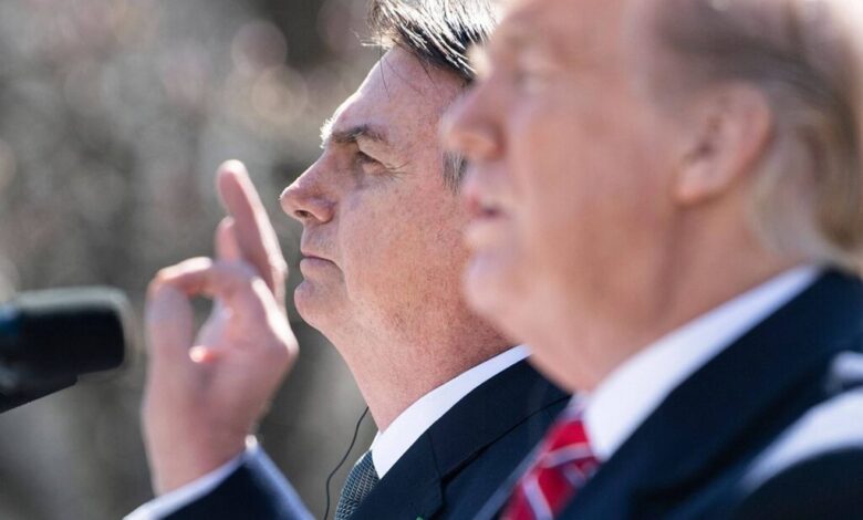 A imagem mostra dois homens em um evento ao ar livre. O homem à esquerda está de perfil, com a mão levantada, enquanto o homem à direita está em foco, mas parcialmente desfocado. Ambos parecem estar em um momento de discurso ou apresentação, com microfones à frente. O fundo é desfocado, sugerindo um ambiente de conferência ou coletiva de imprensa.