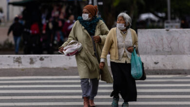 Duas mulheres idosas estão atravessando uma faixa de pedestres. Ambas usam máscaras faciais. A mulher à esquerda está vestida com um casaco longo e um lenço na cabeça, segurando uma sacola. A mulher à direita usa um suéter claro e também carrega uma sacola. Ao fundo, há pessoas e uma estrutura que parece ser um quiosque.