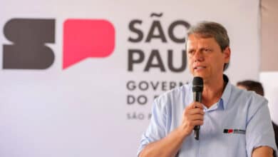 Um homem está falando ao microfone em um evento. Ele usa uma camisa azul clara e está em frente a um fundo com a logo do governo de São Paulo, que inclui as letras