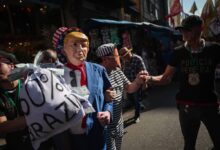 A imagem mostra um grupo de pessoas em um protesto. Um homem está usando uma máscara que se assemelha a um ex-presidente dos Estados Unidos, vestido com um terno azul e segurando um cartaz que diz