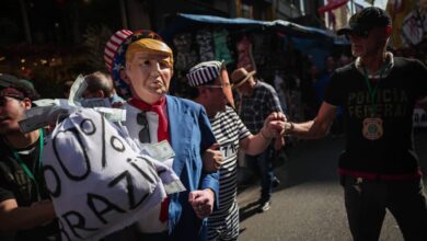 A imagem mostra um grupo de pessoas em um protesto. Um homem está usando uma máscara que se assemelha a um ex-presidente dos Estados Unidos, vestido com um terno azul e segurando um cartaz que diz