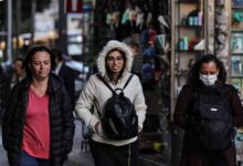 Três mulheres caminham pela calçada em uma área urbana. A mulher à esquerda usa uma jaqueta preta e tem cabelo solto. A mulher do meio usa um casaco claro com capuz e óculos, carregando uma mochila preta. A mulher à direita está usando uma máscara facial e também carrega uma mochila. Ao fundo, há uma vitrine com diversos objetos expostos e outras pessoas caminhando.