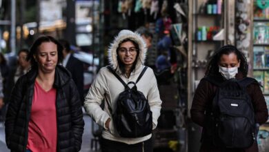 Três mulheres caminham pela calçada em uma área urbana. A mulher à esquerda usa uma jaqueta preta e tem cabelo solto. A mulher do meio usa um casaco claro com capuz e óculos, carregando uma mochila preta. A mulher à direita está usando uma máscara facial e também carrega uma mochila. Ao fundo, há uma vitrine com diversos objetos expostos e outras pessoas caminhando.