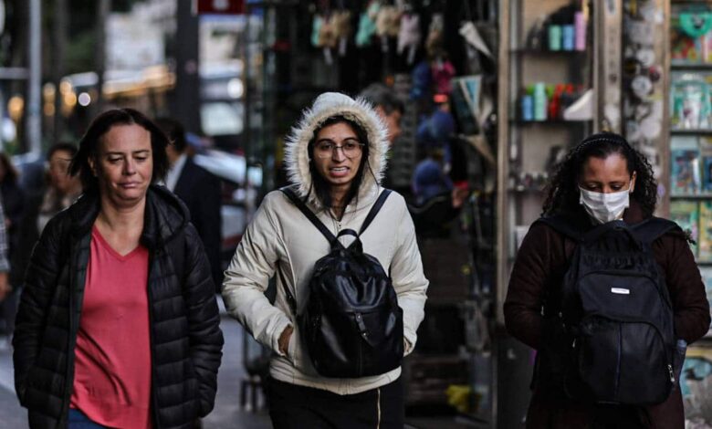 Três mulheres caminham pela calçada em uma área urbana. A mulher à esquerda usa uma jaqueta preta e tem cabelo solto. A mulher do meio usa um casaco claro com capuz e óculos, carregando uma mochila preta. A mulher à direita está usando uma máscara facial e também carrega uma mochila. Ao fundo, há uma vitrine com diversos objetos expostos e outras pessoas caminhando.