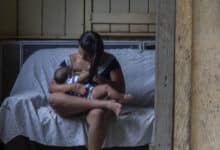 A imagem mostra uma mulher sentada em um sofá, amamentando um bebê. Ela está vestida com uma blusa listrada e calças. Ao fundo, uma outra mulher está de pé, olhando para a cena através de um espelho. A parede é de madeira e o ambiente parece ser uma casa simples, com janelas que mostram vegetação do lado de fora.