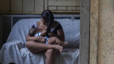 A imagem mostra uma mulher sentada em um sofá, amamentando um bebê. Ela está vestida com uma blusa listrada e calças. Ao fundo, uma outra mulher está de pé, olhando para a cena através de um espelho. A parede é de madeira e o ambiente parece ser uma casa simples, com janelas que mostram vegetação do lado de fora.