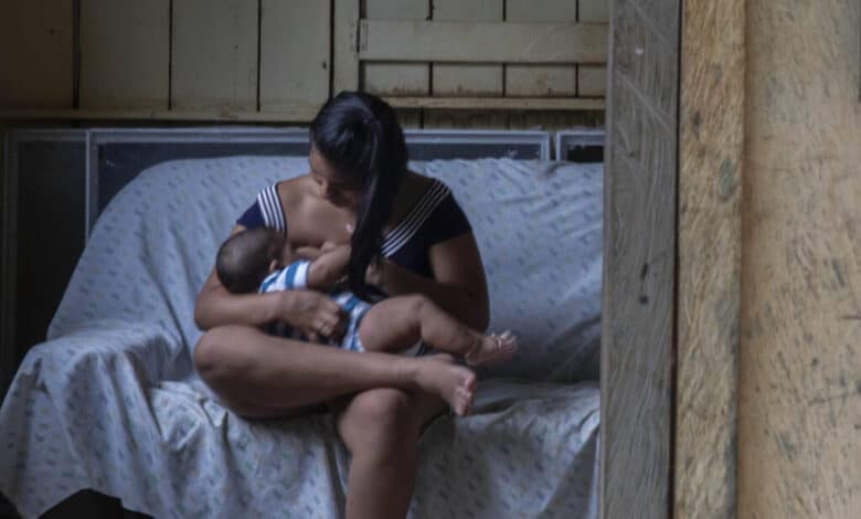 A imagem mostra uma mulher sentada em um sofá, amamentando um bebê. Ela está vestida com uma blusa listrada e calças. Ao fundo, uma outra mulher está de pé, olhando para a cena através de um espelho. A parede é de madeira e o ambiente parece ser uma casa simples, com janelas que mostram vegetação do lado de fora.