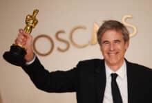 Um homem sorridente está segurando um troféu do Oscar com uma mão levantada. Ele está vestido com um terno preto e uma gravata preta, e ao fundo há uma parede com a palavra