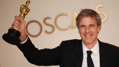 Um homem sorridente está segurando um troféu do Oscar com uma mão levantada. Ele está vestido com um terno preto e uma gravata preta, e ao fundo há uma parede com a palavra