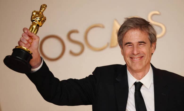Um homem sorridente está segurando um troféu do Oscar com uma mão levantada. Ele está vestido com um terno preto e uma gravata preta, e ao fundo há uma parede com a palavra