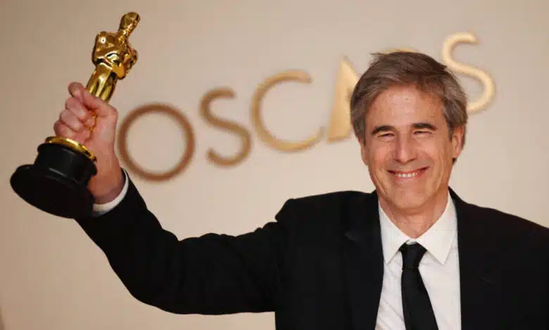 Um homem sorridente está segurando um troféu do Oscar com uma mão levantada. Ele está vestido com um terno preto e uma gravata preta, e ao fundo há uma parede com a palavra