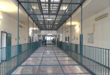 A imagem mostra um corredor de prisão com paredes brancas e um piso de metal. O corredor é cercado por grades metálicas e possui portas em ambos os lados. Há iluminação artificial nas paredes e uma grade no teto, permitindo a entrada de luz natural. O ambiente parece limpo e organizado, com um espaço amplo e sem pessoas visíveis.