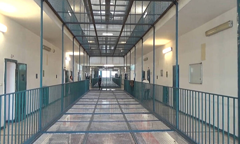 A imagem mostra um corredor de prisão com paredes brancas e um piso de metal. O corredor é cercado por grades metálicas e possui portas em ambos os lados. Há iluminação artificial nas paredes e uma grade no teto, permitindo a entrada de luz natural. O ambiente parece limpo e organizado, com um espaço amplo e sem pessoas visíveis.
