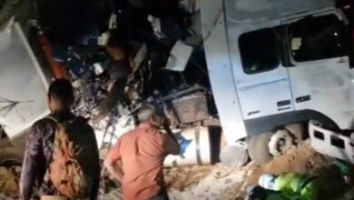 A imagem mostra um caminhão acidentado, com a cabine e a parte traseira danificadas. Há pessoas em pé, observando a cena, e alguns equipamentos de resgate visíveis no chão. O ambiente parece ser noturno, com pouca iluminação.