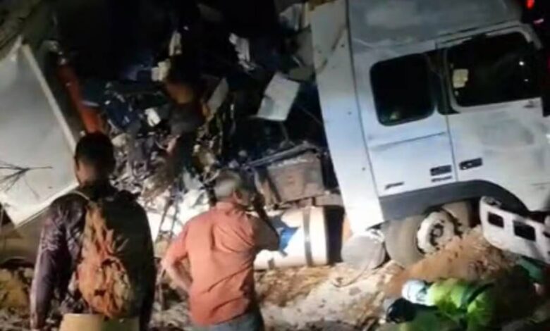 A imagem mostra um caminhão acidentado, com a cabine e a parte traseira danificadas. Há pessoas em pé, observando a cena, e alguns equipamentos de resgate visíveis no chão. O ambiente parece ser noturno, com pouca iluminação.
